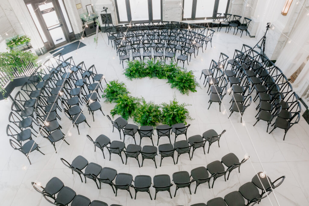 Indoor wedding ceremony with circular greenery in the middle and black chairs surrounding the alter.