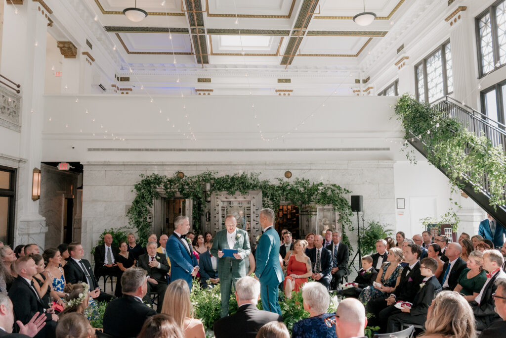 guests seated in circular seatings wearing formal attire facing 2 grooms in blue tuxes, and an officiant in a gray suit