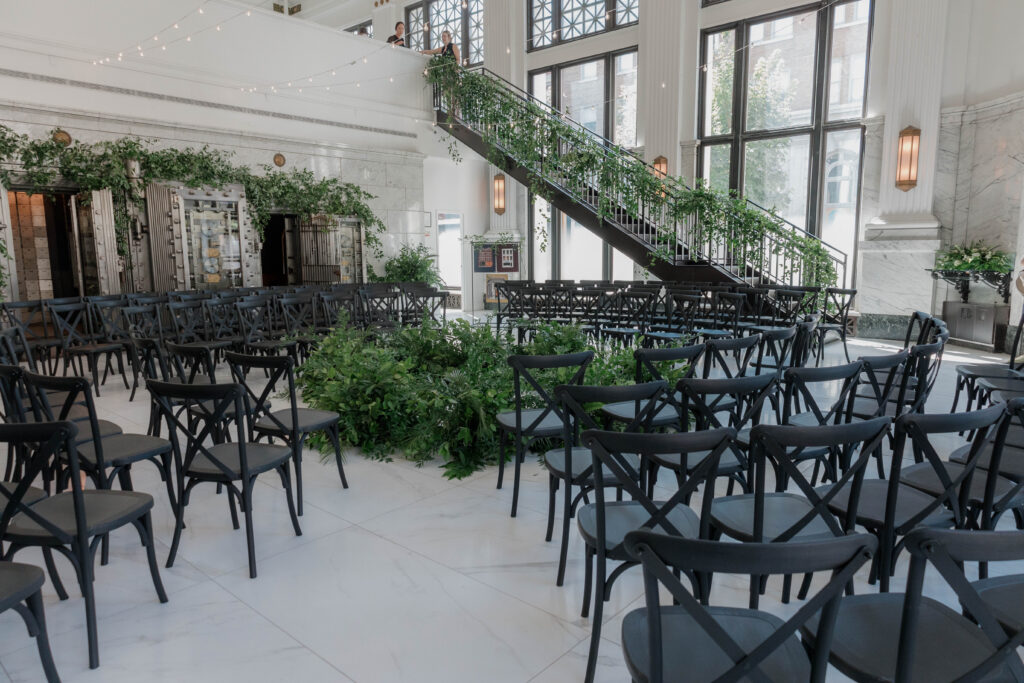 Image of indoor wedding ceremony with circular greenery ground arrangements and black chairs surrounding the alter.