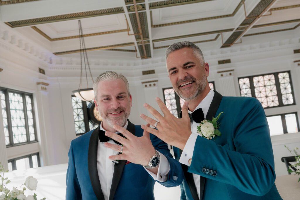 two grooms holding up their left hands with their wedding bands on.