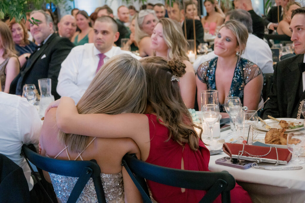 young girl in a red dress hugging a woman in a silver dress while sitting at a wedding reception.