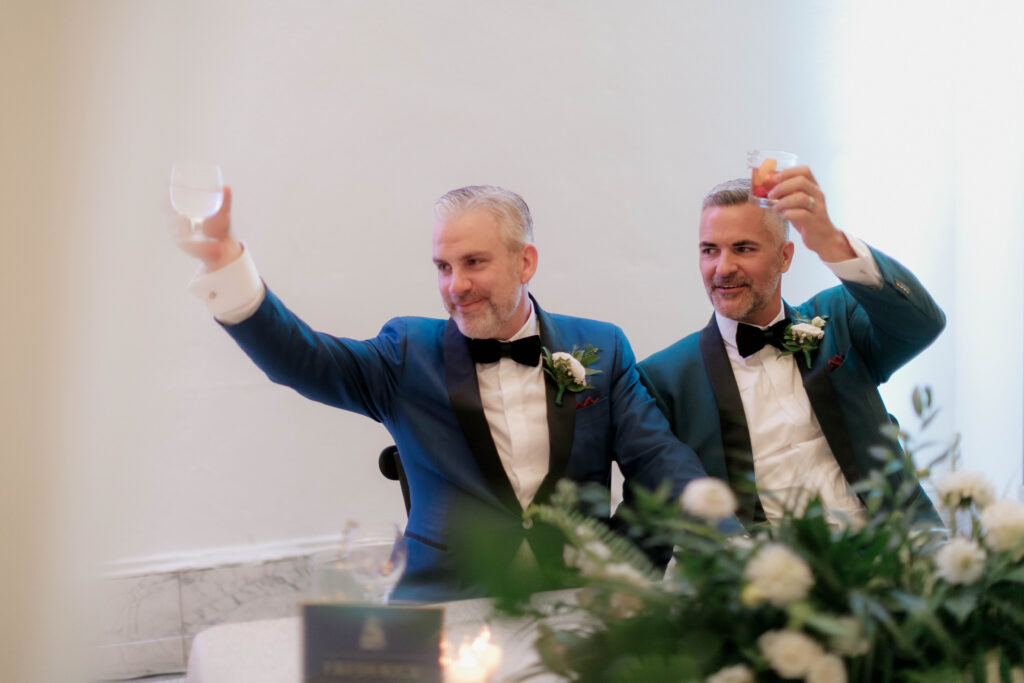 two grooms in blue tuxes holding glasses up in a toast