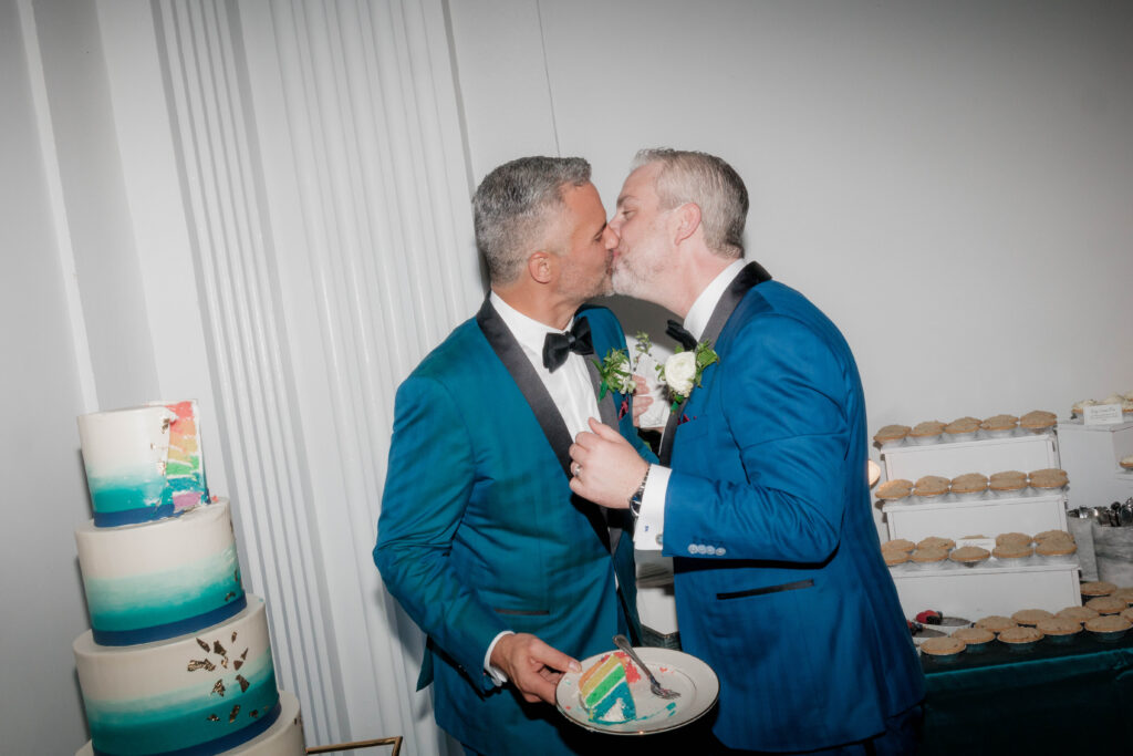two men in blue tuxes kissing while holding a slice of rainbow cake next to a blue tiered wedding cake