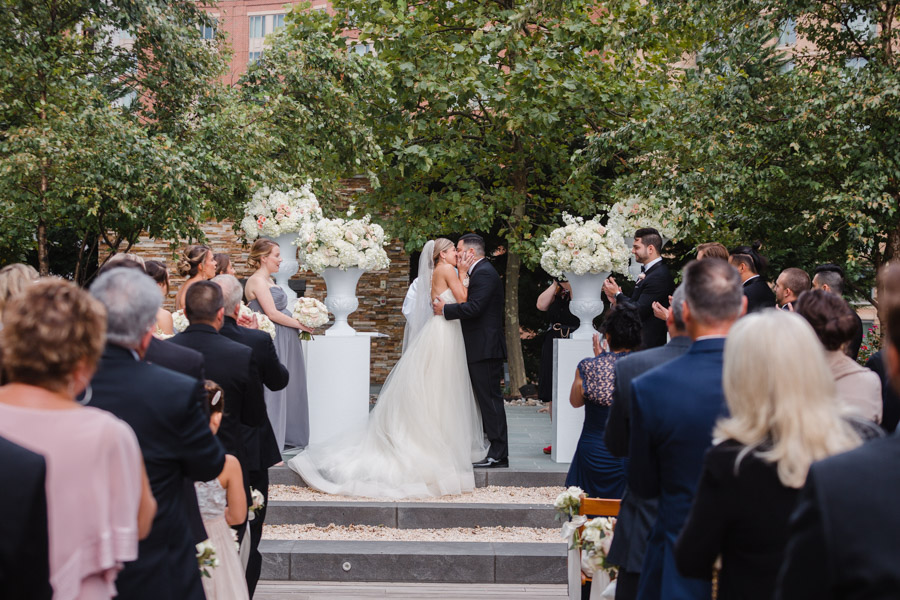 Beautiful bride and groom kissing at the end of the aisle, flanked by trees and white urns filled with white flowers. The guests are all standing.