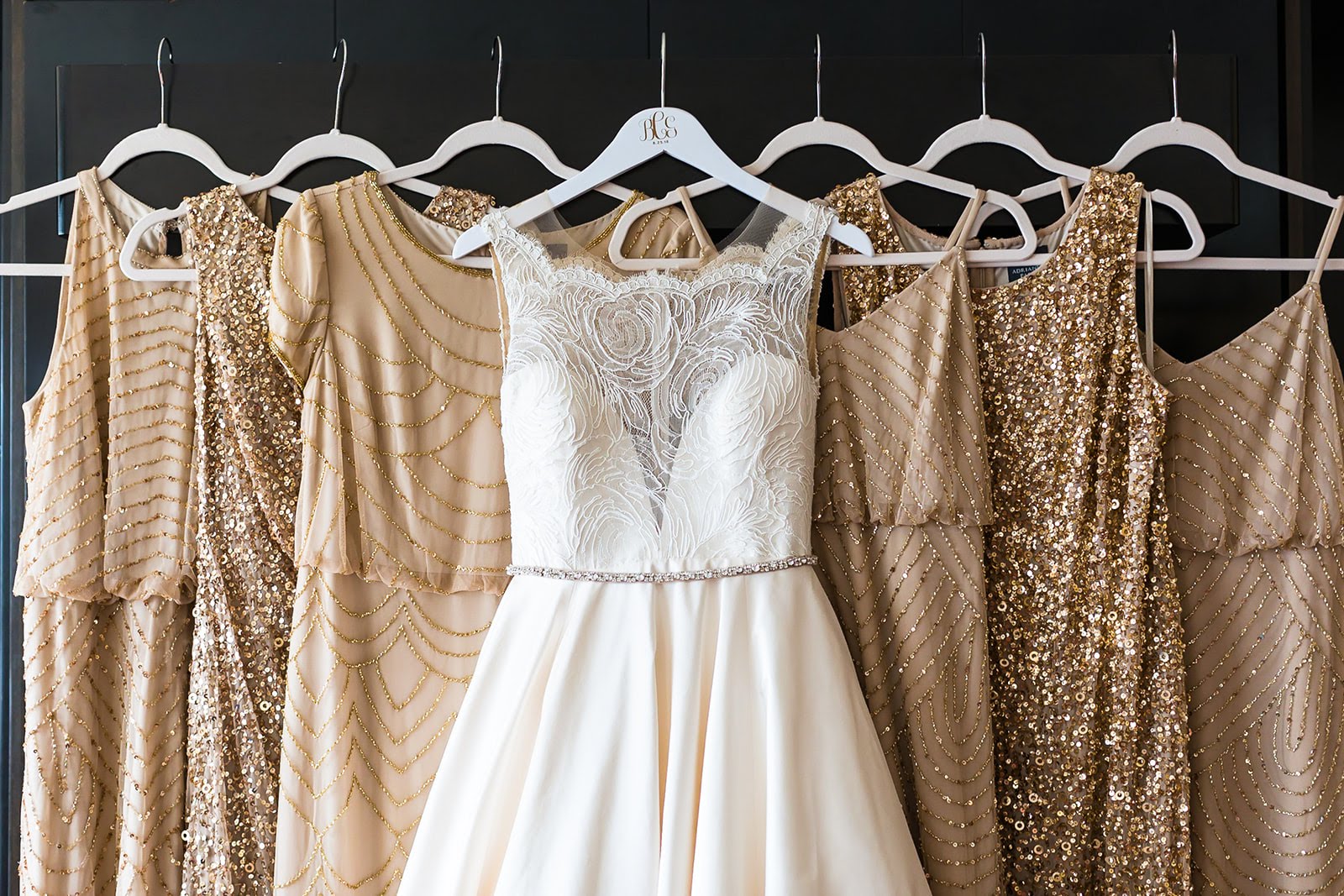 Czerwinkski-Getting-Ready-Bride-30 (1) A white bridal gown hangs in the center, flanked by six mismatched bridesmaid dresses in shades of gold and rose gold with varying textures and levels of sparkle.