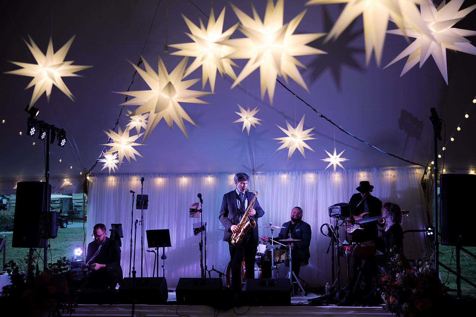 Laura & Shawn's Wedding A band performs on a stage under a tent illuminated by multiple large, star-shaped paper lanterns and string lights. The band members are playing instruments, including a saxophone.
