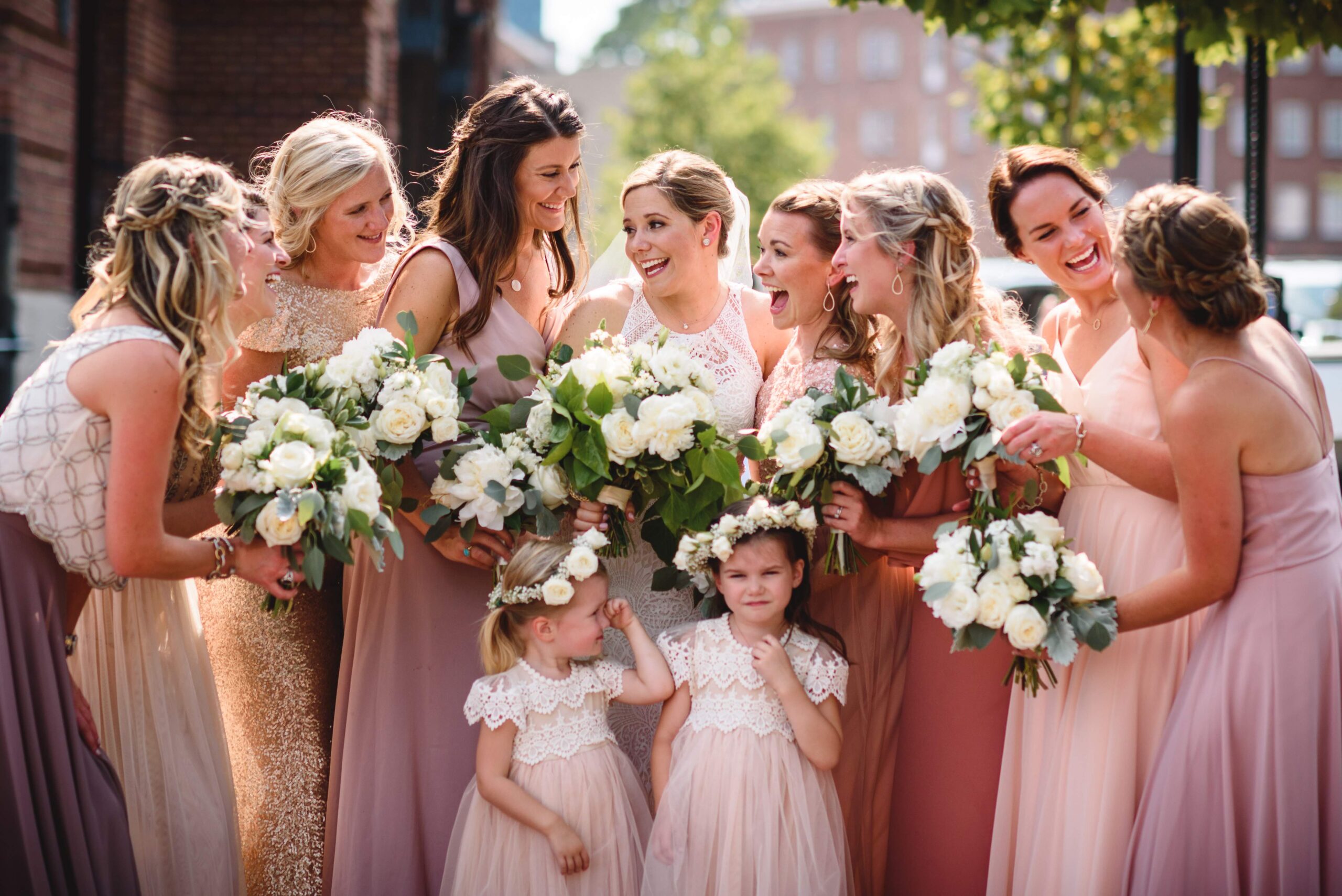 julcolwed_weddingparty20 A group of bridesmaids and two flower girls, all in varying shades of pink and gold dresses, smiling and holding bouquets.